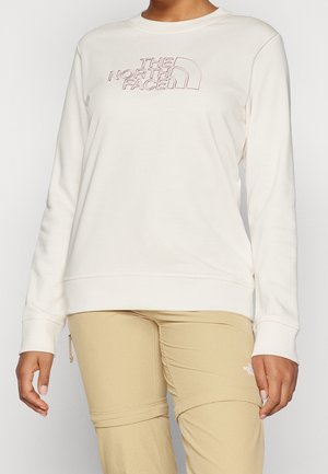 Personne portant un sweat-shirt blanc à manches longues avec le logo The North Face et un pantalon beige avec des coutures visibles et des poches zippées.