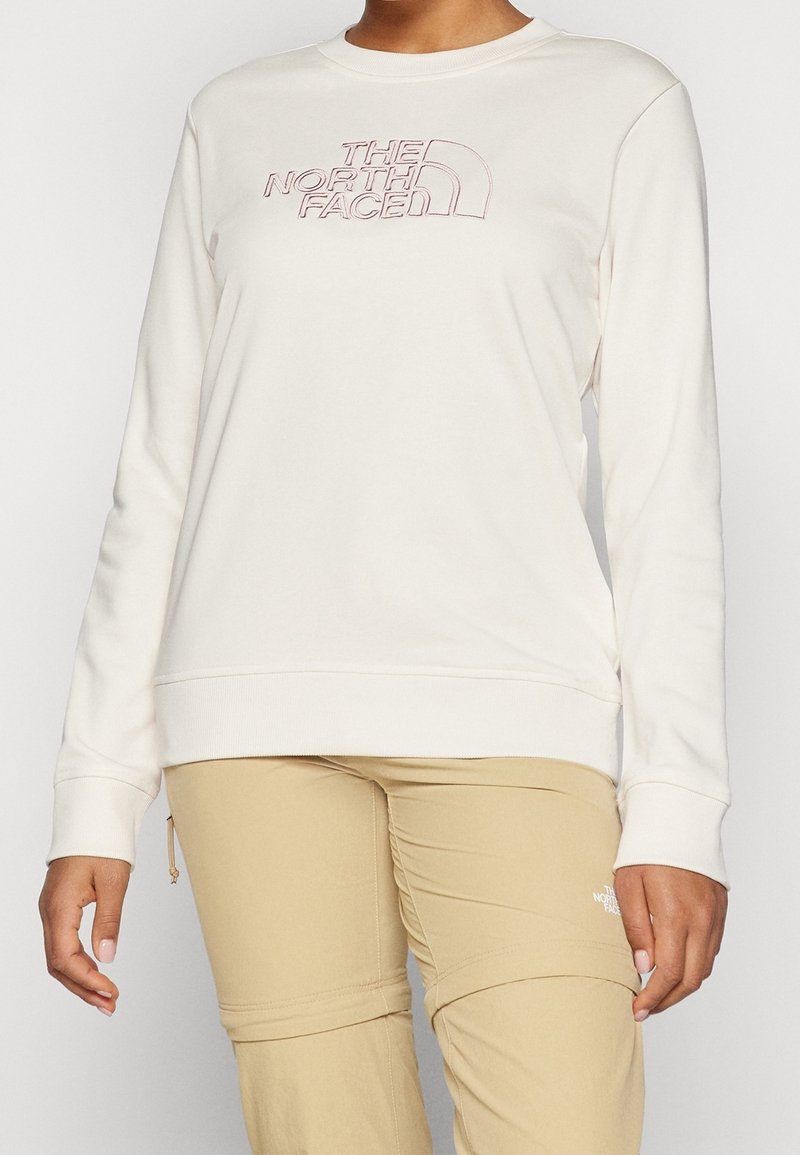 Personne portant un sweat-shirt blanc à manches longues avec le logo The North Face et un pantalon beige avec des coutures visibles et des poches zippées.