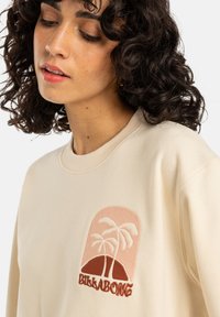 Crème sweatshirt met een ronde graphic van roze en bruine palmbomen en een bergontwerp, naast geborduurde "Billabong" tekst.