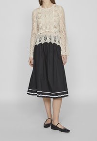 Top in cotone lavorato all'uncinetto di colore crema con maniche lunghe abbinato a una gonna nera a ginocchio con un motivo a zigzag bianco lungo l'orlo. Scarpe nere basse.