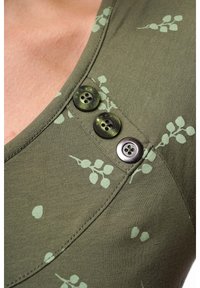 Grünes Strickoberteil mit Blättermustern, einem tiefen Ausschnitt und drei Knöpfen – zwei in Grün und einer in Dunkelbraun – auf der linken Seite.