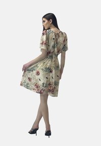 Vestido floral con mangas cortas, cintura ajustada, tejido color beige claro, que presenta patrones florales rojos y verdes, y un detalle de abertura en la espalda.