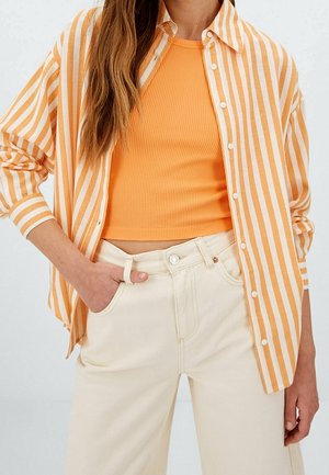 Person iført en gul-orange ribbet crop top, cremet højtaljede bukser og en åben gul-hvid stribet skjorte med hånden i lommen.