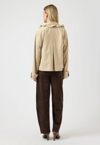 Vrouw met beige jas met gerafelde kraag, bruine broek met wijde pijpen en beige schoenen met hakken, staand met haar rug naar de camera tegen een effen achtergrond.