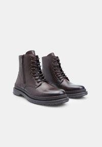 Botas de tobillo de cuero marrón con cordones en la parte delantera y cremallera lateral. Presentan una puntera redondeada y una suela de goma texturizada para mayor agarre.