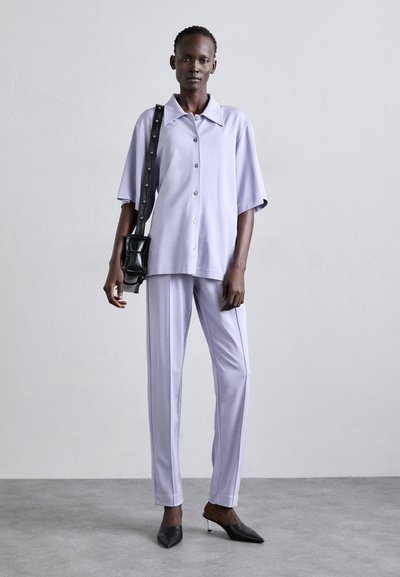 Lilac short-sleeve button-up shirt paired with matching tailored pants. Black crossbody bag and pointed black heels complete the outfit.