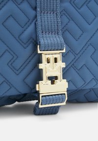 Niebieska pikowana torba z tkaniny z wyraźną złotą klamrą i plecionym paskiem, prezentująca geometryczny wzór i teksturowaną powierzchnię.