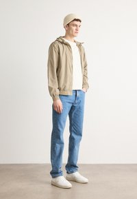 Letthvid beige jakke med elastisk manchetter, båret over en cremefarvet skjorte. Parret med blå jeans og hvide sneakers, toppet med en cremefarvet strikket beanie.