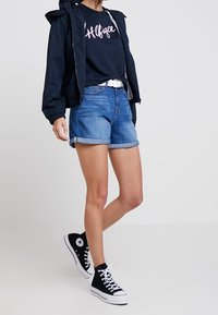 Granatowy wiatrówka z kapturem, różowa koszulka z logo, dżinsowe szorty i czarne trampki za kostkę z białymi akcentami.