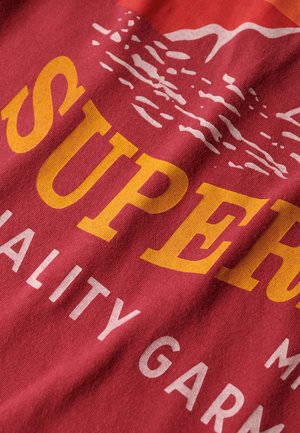 Tessuto di cotone rosso con testo giallo e bianco in evidenza che recita "SUPER" e "QUALITY GARMENT", con un grafico di montagna sopra.