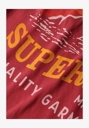 Roter Baumwollstoff mit auffälligem gelben und weißen Text, der "SUPER" und "QUALITÄTSGEWAND" zeigt, mit einer Berggrafik darüber.