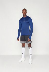 Kék hosszú ujjú sportfelső, szürke rövidnadrág két fekete csíkkal, fehér zokni fekete díszítéssel és fehér stoplis cipő, bábon bemutatva.