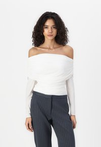 Top blanco sin mangas con drapeado texturizado, superpuesto sobre mangas largas. Combinado con pantalones de rayas finas en azul marino oscuro.