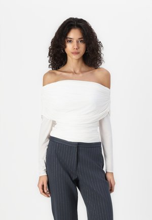 Off-the-shoulder white top with textured draping, layered over long sleeves. Paired with dark navy pinstripe trousers.