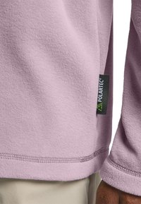 Lila Fleece-Pullover mit weichem Textil, sichtbaren Nähten am Saum und an den Bündchen sowie einem schwarzen Polartec®-Label an der Seite.