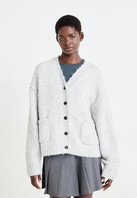 Cardigan lavorato a maglia grigio chiaro con scollo a V, chiusura centrale con bottoni, due tasche frontali e polsini e orlo a costine. Indossato sopra una maglia scura.