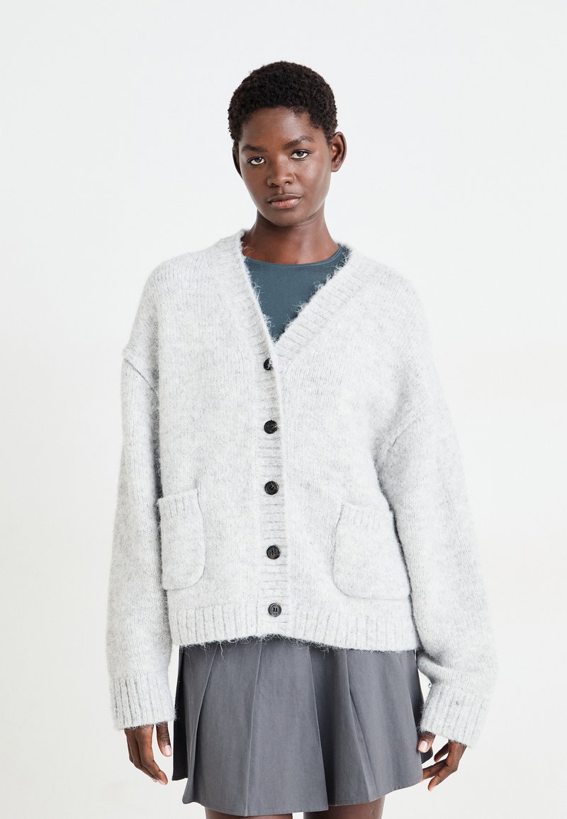 Cardigan lavorato a maglia grigio chiaro con scollo a V, chiusura centrale con bottoni, due tasche frontali e polsini e orlo a costine. Indossato sopra una maglia scura.