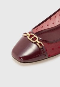 Burgundy patent leather flat with a pointed toe, featuring a sheer dotted fabric panel and a gold chain accent on the front.