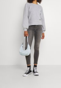 Kvinna som bär grå tröja, grå skinny jeans, svarta högklackade sneakers, håller en ljusblå handväska vid sidan mot vit bakgrund.