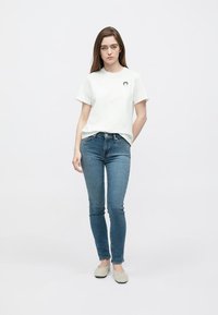 White cotton t-shirt with short sleeves and a round neckline, featuring a small black graphic. Paired with dark blue skinny jeans and light shoes.