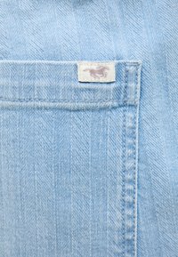 Tela de algodón vaquero de color azul claro que presenta un bolsillo frontal con un borde cosido y una etiqueta tejida que muestra un diseño de caballo. Superficie texturizada visible.