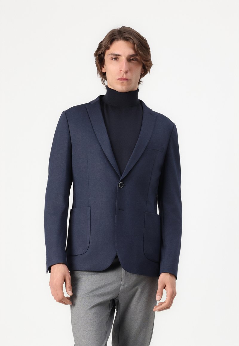 Blazer bleu marine avec revers crantés, fermeture à un bouton et deux poches avant. Porté par-dessus un col roulé noir. Texture fluide.
