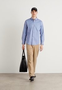 Camisa de botão azul claro, calças bege, sapatos pretos, a segurar uma mala de mão preta, de pé contra um fundo branco liso.