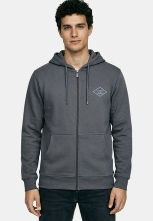 Jeune homme aux cheveux bouclés portant un sweat à capuche gris avec fermeture éclair, poches avant et un petit logo sur la poitrine, debout devant un fond uni.