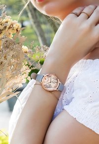Reloj redondo con caja de oro rosa y correa de silicio gris claro. La esfera presenta diseños florales y marcadores de horas. Lleva puesto en una muñeca con anillos.