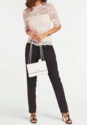 Femme portant un haut en dentelle rose pâle et un pantalon noir à motifs, tenant un sac à main Guess blanc à bandoulière chaîne, chaussée de talons noirs à brides.