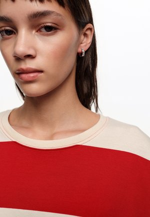 Persona joven con cabello corto y oscuro, que lleva pendientes de aro plateados y una camiseta de cuello redondo en tonos rojo y beige, sobre un fondo blanco.