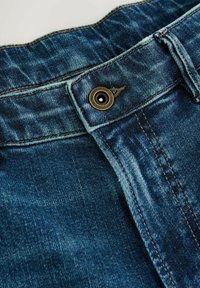 Denimová tkanina v tmavomodrej farbe s vyblednutou textúrou. Obsahuje kovový gombík s vyrytou nálepkou "Prémiová kvalita" a kontrastným prešívaním.