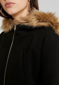 Sort hættefrakke med en kunstpelskrave, der har en sølvlynlås og sidelomme, der fremviser et blødt, textureret stof.