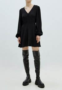 Zwarte lange mouwen jurk met een V-hals, geplooide taille en knopen. Gepaard met kniehoge zwarte leren laarzen met stevige zolen.