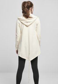 Sudadera color crema con un dobladillo ajustable, mangas largas con cremalleras y un fondo asimétrico. Tejido suave con una textura lisa.