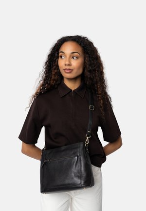 Bolso de hombro de piel negra con un bolsillo frontal con cremallera y detalles en hardware dorado, combinado con una camisa polo marrón y pantalones de color claro.