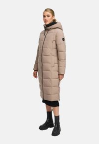 Abrigo puffer largo beige con capucha, diseño acolchado y bolsillos laterales. Cierre frontal con cremallera y acentos negros. Llevado sobre un suéter negro y botas.
