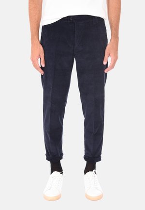 Mann trägt marineblaue Cordhosen mit hochgekrempelten Säumen, weiße Sneakers, schwarze Socken und ein weißes Hemd, steht vor einem schlichten Hintergrund.