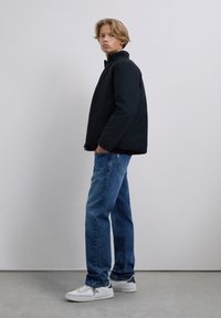 Junge Person mit blonden Haaren, die eine schwarze Jacke, blaue Jeans und weiße Sneakers trägt, steht vor einem schlichten hellgrauen Hintergrund.