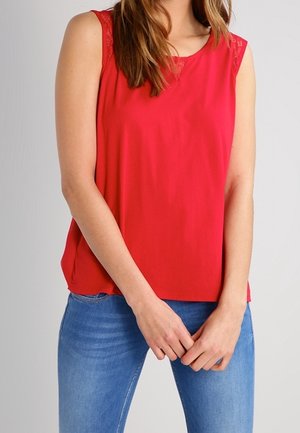 Rød ærmeløs bluse med blondedetalje på skuldrene, båret med blå jeans, modellen står med hænderne løst foldet foran.