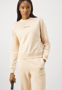 Felpa beige chiaro con collo rotondo e maniche lunghe. Presenta la scritta "BJÖRN BORG" ricamata in verde. Abbinata a pantaloni della tuta coordinati.