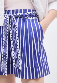 Κοντινή λήψη μπλε και λευκών κάθετων ριγέ σορτς με ταιριαστή ζώνη δεμένη σε φιόγκο, φορεμένα με ανοιχτόχρωμο μπουφάν και χέρι στην τσέπη.