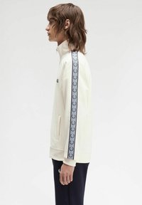 Chaqueta cremosa con cremallera y cuello alto, con cinta de logo en azul marino a lo largo de las mangas. Presenta bolsillos laterales y puños de canalé. Combinada con pantalones azul marino.