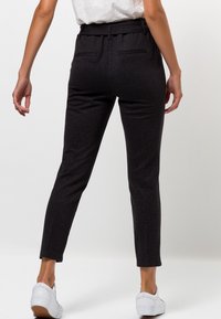 Pantalon noir ajusté en tissu texturé, taille mi-haute, ceinture élastique, avec deux poches arrière, porté avec des baskets blanches.
