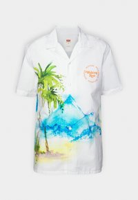 Camisa blanca de manga corta con un diseño acuarela de palmeras y montañas en verde y azul. Incluye un notable detalle de texto circular.