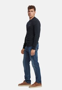 Suéter de punto texturizado azul marino con cuello redondo, combinado con jeans azules y zapatillas de ante marrón. Logo pequeño destacado en el pecho del suéter.
