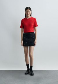 Νεαρή γυναίκα που φοράει κόκκινο κοντομάνικο τοπ και μαύρη μίνι φούστα με μικρά λευκά σχέδια, μαύρες κάλτσες και μαύρα μοκασίνια, μπροστά σε γκρίζο τοίχο.