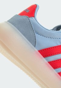 Ljusblå sneakers med röda läderaccenter och tre röda ränder. Har texturerad grå mocka och en genomskinlig gummisula.