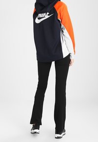 Felpa con cappuccio blu navy, arancione e bianco, con un grande logo Nike bianco sulla schiena. Abbinata a pantaloni neri a zampa d'elefante e scarpe da ginnastica.