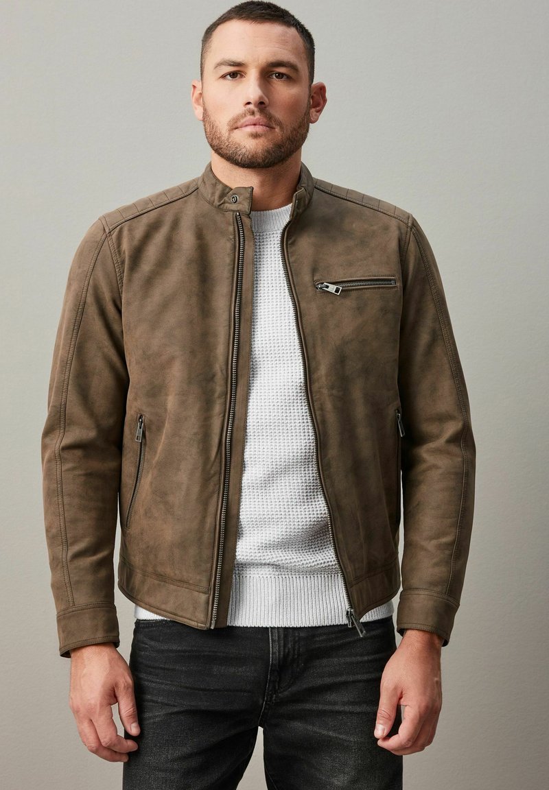 Chaqueta de cuero sintético marrón con cuello alzado, bolsillos con cremallera en los laterales y el pecho, y detalles texturizados en los hombros, llevada sobre un suéter de punto blanco.